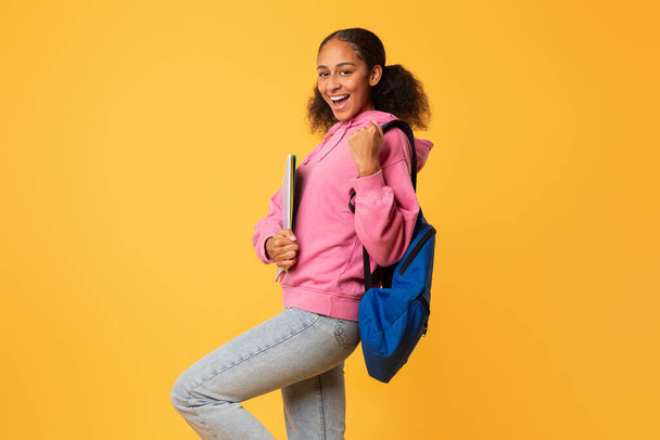 Happy black teen student lady gestikuliert ja mit College-Materialien und trägt Rucksack, auf gelbem Studiohintergrund mit freiem Platz, feiert akademischen Erfolg am College - Foto, Bild