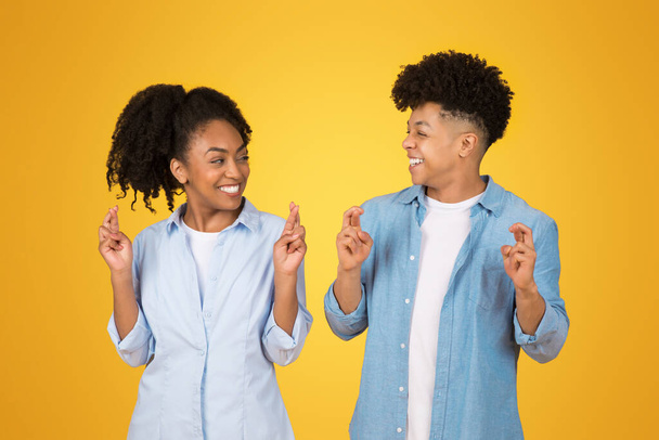 Optimiste heureux jeunes frères et sœurs afro-américains en costume bleu clair faire des gestes de souhait, souriant et espérant avec les doigts croisés sur un fond jaune vif, studio - Photo, image