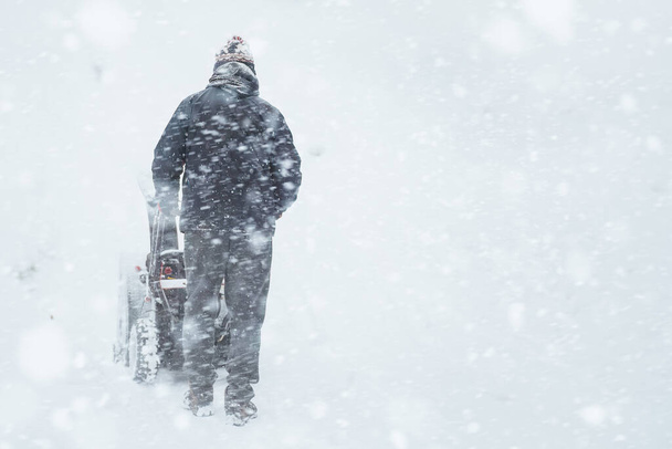 Una escena suburbana nevada con un hombre usando un soplador de nieve para despejar la acera. Un hombre limpia la nieve de la calle con un potente soplador de nieve después de una fuerte tormenta de invierno. Temporada de invierno trabajo fuera. - Foto, imagen