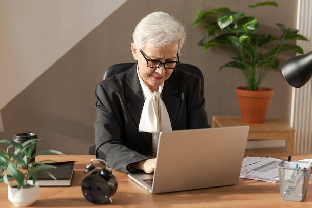 Confident stylish european middle aged senior woman using laptop at workplace. Stylish older mature 60s gray haired lady businesswoman sitting at office table. Boss leader teacher professional worker - Foto, afbeelding