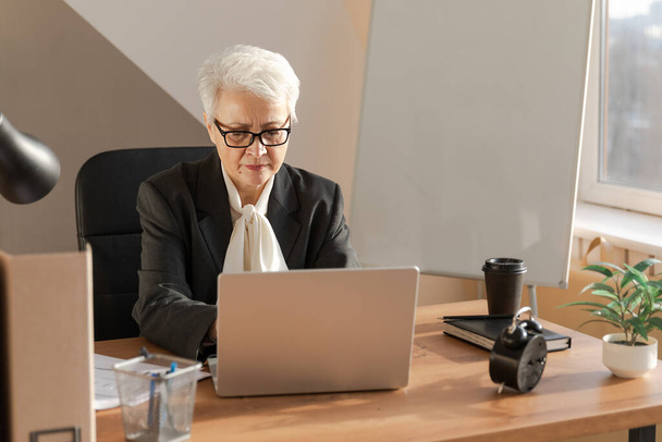 Confident stylish european middle aged senior woman using laptop at workplace. Stylish older mature 60s gray haired lady businesswoman sitting at office table. Boss leader teacher professional worker - Foto, immagini