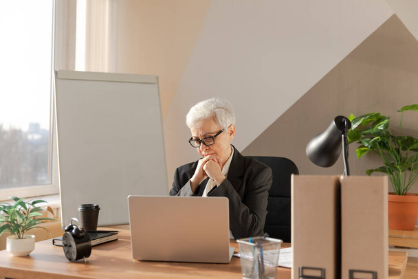 Confident stylish european middle aged senior woman using laptop at workplace. Stylish older mature 60s gray haired lady businesswoman sitting at office table. Boss leader teacher professional worker - Foto, afbeelding