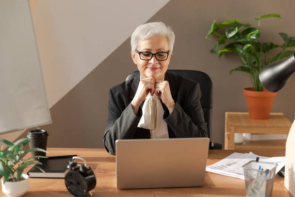 Уверенная стильная европейская старшая женщина средних лет с ноутбуком на рабочем месте. Стильная взрослая женщина 60-х годов, седовласая деловая женщина, сидящая за рабочим столом. Профессиональный работник - Фото, изображение