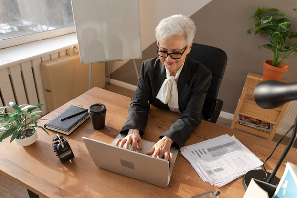 Αυτοπεποίθηση stylish Ευρωπαίος μεσήλικας ηλικιωμένη γυναίκα χρησιμοποιώντας φορητό υπολογιστή στο χώρο εργασίας. Κομψή μεγαλύτερη ώριμη 60άρα γυναίκα επιχειρηματίας με γκρίζα μαλλιά κάθεται στο τραπέζι του γραφείου. Αρχηγός εκπαιδευτικός επαγγελματίας εργαζόμενος - Φωτογραφία, εικόνα