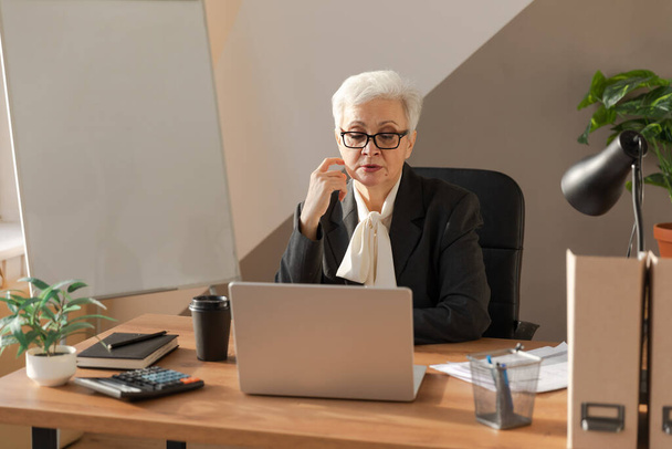 İş yerinde dizüstü bilgisayar kullanan kendine güvenen Avrupalı orta yaşlı bir kadın. Modaya uygun, olgun 60 'ların beyaz saçlı iş kadını ofis masasında oturuyor. Patron öğretmen profesyonel işçi. - Fotoğraf, Görsel