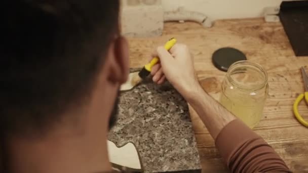 Над плечом снимок сапожника мужчина нанося клей с кистью на кожаный кусок, сидя за столом в мастерской - Кадры, видео