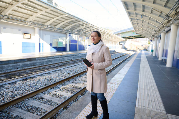 Teljes hosszúságú portré női ingázó kezében okos mobiltelefon, várja, hogy felszálljon a vonatra, állt a peronon egy vasútállomás, a kék ég háttér télen. Emberek! Életmód - Fotó, kép