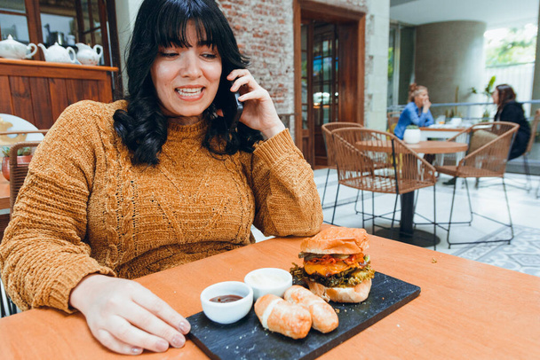 молодая женщина из Латинской Венесуэлы, сидящая в ресторане, расстроена разговором по телефону со своей подругой, которая не пришла вовремя на обед, с тарелкой гамбургера и текино с соусом на столе. - Фото, изображение