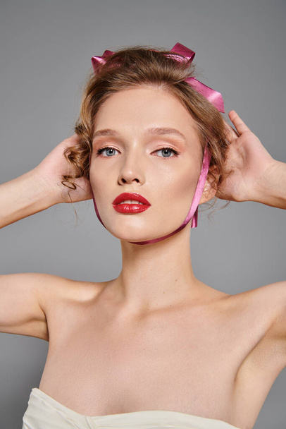 Młoda kobieta z klasycznym pięknem uderza w pozę w studio, nosząc różowy łuk na głowie na szarym tle. - Zdjęcie, obraz