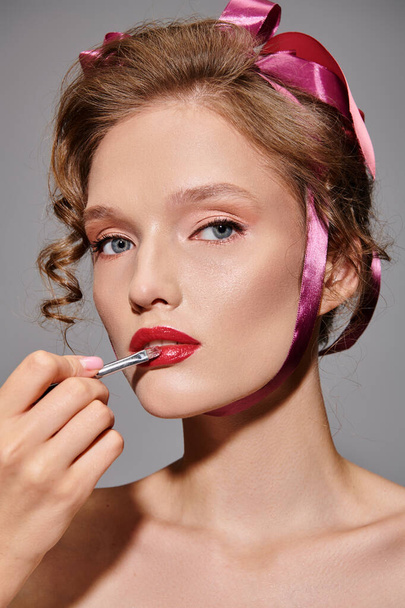 Een jonge vrouw met een roze strik in haar haar brengt lippenstift aan op haar lippen, straalt klassieke schoonheid en elegantie uit in een studio op een grijze achtergrond. - Foto, afbeelding