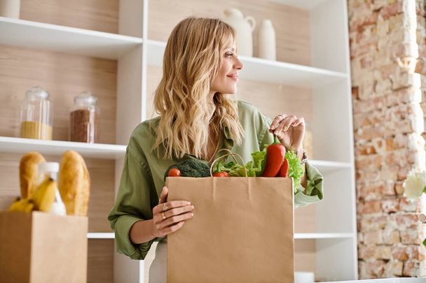 Una mujer de pie en una cocina, sosteniendo una bolsa llena de verduras. - Foto, Imagen