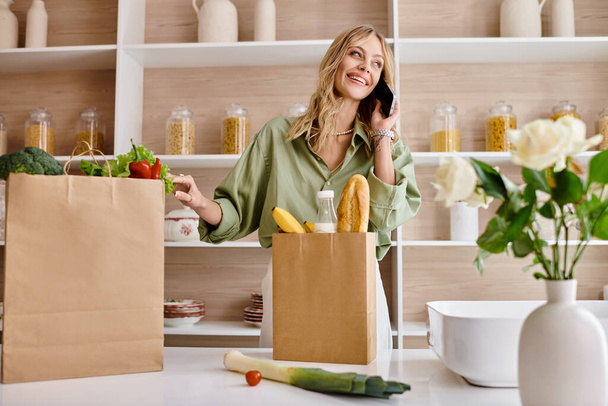 Femme multitâche avec téléphone portable et sac à provisions dans la cuisine. - Photo, image
