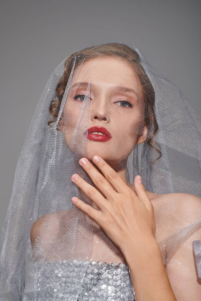 A young woman exuding classic beauty, wearing a veil and vibrant red lipstick in a studio setting on a grey background. - Photo, Image