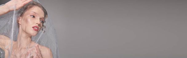 Een jonge vrouw met een sluier die haar haar sierlijk in de lucht houdt en klassieke schoonheid uitstraalt in een studio op een grijze achtergrond. - Foto, afbeelding
