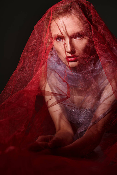 A young woman exudes classic beauty in a stunning red veil and dress, striking a pose in a studio setting against a black backdrop. - 写真・画像
