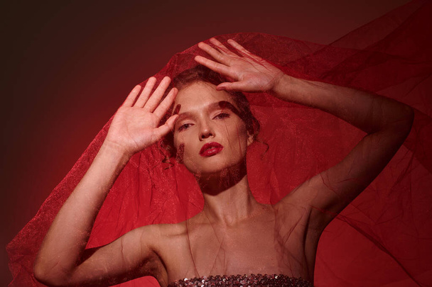 Una giovane donna trasuda bellezza classica, vestita con un vibrante abito rosso con le mani elegantemente posizionate sulla testa. - Foto, immagini