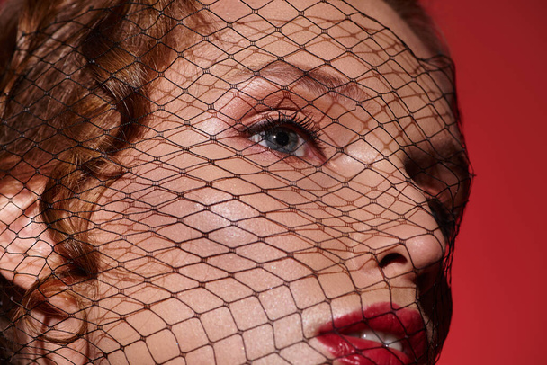 Mladá žena vyzařuje klasickou krásu s červeným závojem zakrývajícím její tvář, vytvářejícím atmosféru tajemna a elegance. - Fotografie, Obrázek