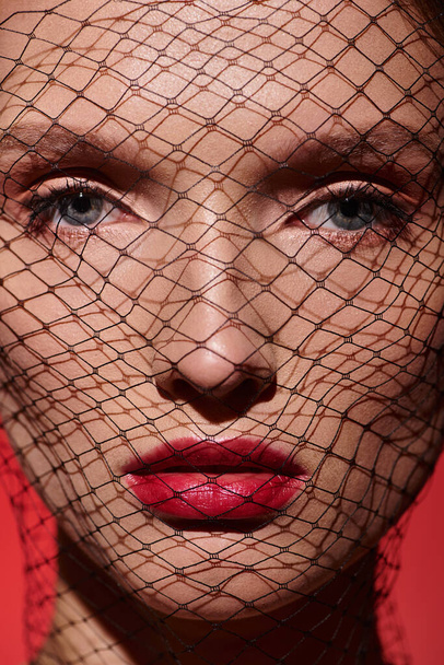 Eine junge Frau mit klassischer Schönheit posiert in einem Studio mit einem roten Schleier, der ihr Gesicht verhüllt und Geheimnisse und Eleganz ausstrahlt. - Foto, Bild