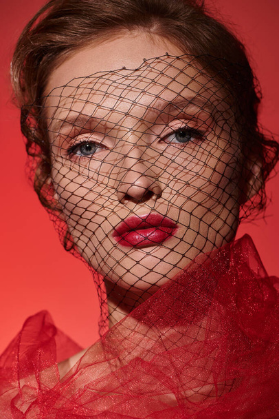 Uma jovem exala a beleza clássica enquanto posa em um estúdio com um véu vermelho cobrindo seu rosto. - Foto, Imagem