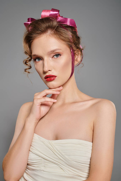 Egy fiatal nő klasszikus szépséget áraszt fehér ruhában, rózsaszín masnival a fején, miközben elegánsan pózol egy stúdióban.. - Fotó, kép