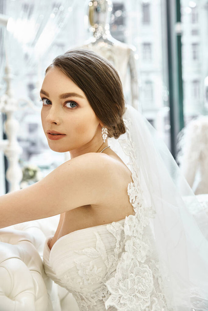A young brunette bride in a flowing white wedding dress, seated gracefully on a luxurious couch in a bridal salon. - Valokuva, kuva