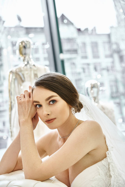 Eine junge brünette Braut in einem fließenden weißen Brautkleid lehnt anmutig auf einem luxuriösen Bett in einem Brautsalon. - Foto, Bild