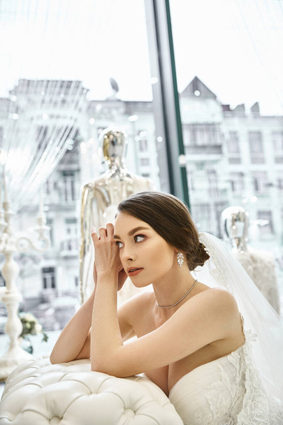 Eine junge brünette Braut in einem wunderschönen Brautkleid sitzt friedlich vor einem Fenster in einem Brautsalon. - Foto, Bild