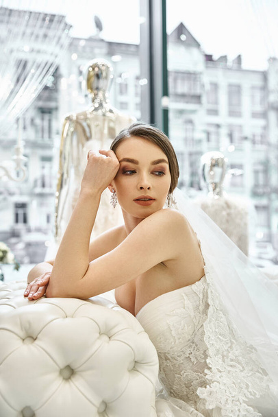 A young brunette bride in a wedding dress gracefully sits on a elegant couch in a bridal salon. - Fotografie, Obrázek