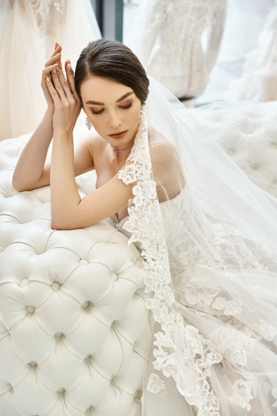 A young brunette bride in a wedding dress lays gracefully on a plush sofa in a luxurious bridal salon. - Foto, Imagem