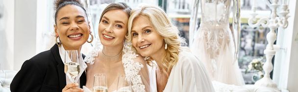 A young brunette bride in a wedding dress and her middle-aged blonde mother in a bridal salon, standing proudly together. - Photo, Image