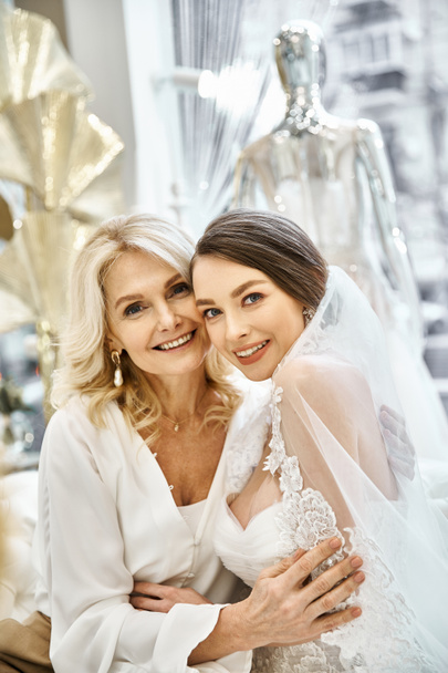 Eine junge brünette Braut im Brautkleid steht neben ihrer blonden Mutter mittleren Alters in einem Brautsalon. - Foto, Bild