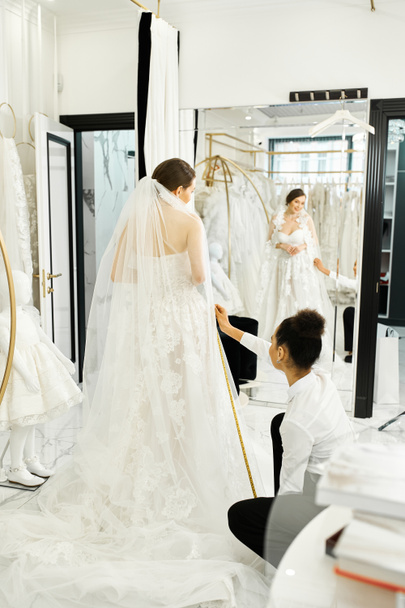 Una joven novia en un vestido de novia blanco conversa con un asistente de compras afroamericano en un lujoso salón nupcial. - Foto, imagen