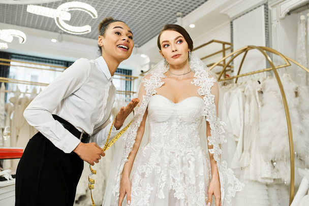 Una joven novia morena en un vestido de novia se encuentra junto a un asistente de compras afroamericano en un salón de novias. - Foto, imagen