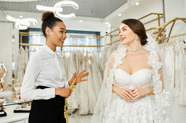 Eine junge brünette Braut im Brautkleid steht neben einer afroamerikanischen Shopping-Assistentin in einem Brautsalon. - Foto, Bild