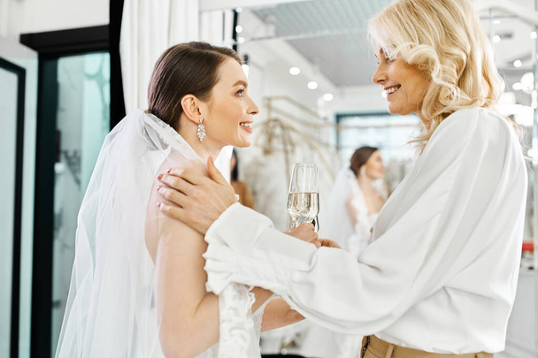 Eine junge Braut im weißen Brautkleid und ihre Mutter, beide elegant in Weiß gekleidet, stoßen mit Champagnergläsern an. - Foto, Bild