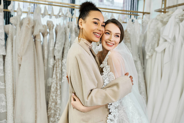 A young bride in a white wedding dress embracing her bridesmaid in front of a rack of various wedding dresses in a bridal salon. - Photo, Image