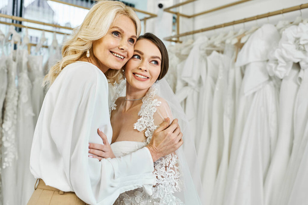 Mladá brunetka nevěsta ve svatebních šatech objímá svou matku středního věku ve svatebním salonu plném svatebních šatů. - Fotografie, Obrázek