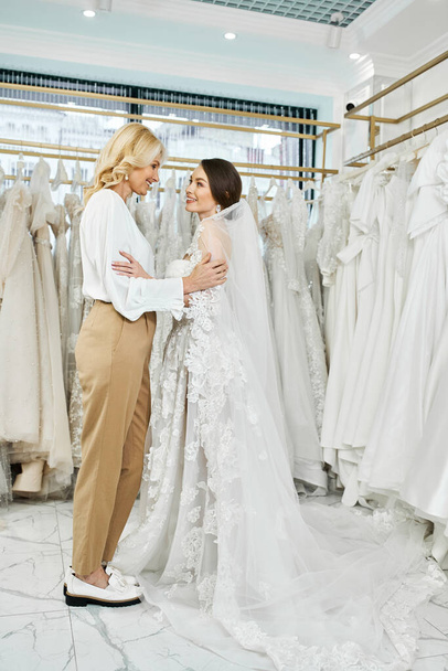 A young brunette bride in a wedding dress and her middle-aged mother stand side by side in a bridal salon, looking at a rack of gowns. - Photo, Image