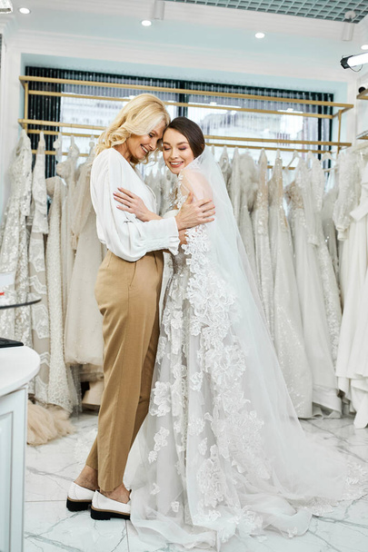 A young bride in a white wedding dress hugs her middle-aged mother, both surrounded by a display of elegant wedding gowns. - 写真・画像