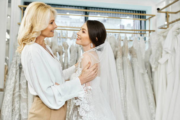 Egy fiatal barna menyasszony és középkorú anyja egymás mellett állnak egy ruhafogas előtt egy menyasszonyi szalonban.. - Fotó, kép