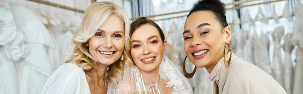 Eine junge Braut im Brautkleid, ihre Mutter mittleren Alters und ihre beste Freundin als Brautjungfer stehen gemeinsam in einem Brautsalon. - Foto, Bild