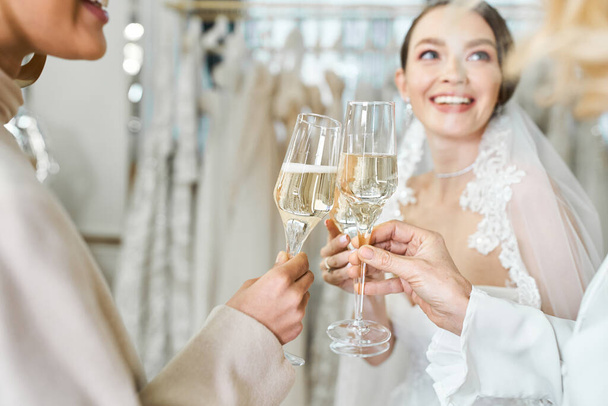 Young bride, her mother, and best friend standing in a bridal salon, raising glasses of champagne in a celebratory toast. - Photo, Image