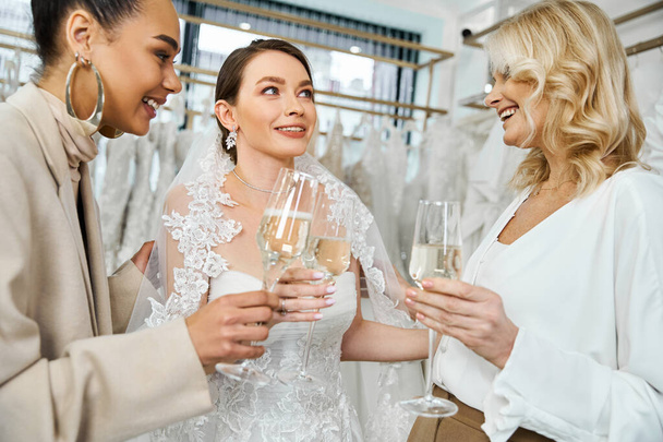 A young brunette bride in a wedding dress, her middle-aged mother, and best friend as a bridesmaid, standing together, holding wine glasses. - Photo, Image