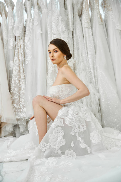 Eine junge brünette Braut in einem weißen Hochzeitskleid sitzt anmutig auf einem Bett und betrachtet den bevorstehenden bedeutsamen Anlass.. - Foto, Bild