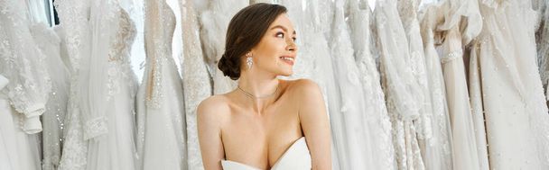 Молодая, красивая брюнетка невеста стоит среди полки белых свадебных платьев в свадебном салоне. - Фото, изображение