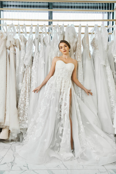 Eine junge brünette Frau, die in einem Hochzeitssalon von einem Gestell eleganter Kleider umgeben ist und über ihre Möglichkeiten für den großen Tag nachdenkt. - Foto, Bild