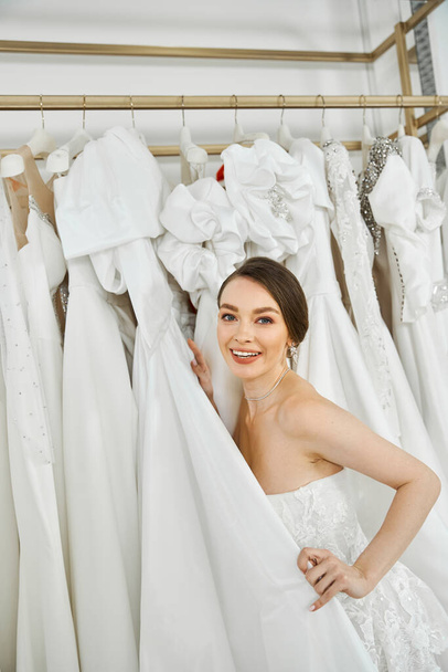 Güzel, genç bir gelin, dalgalı beyaz elbiseli esmer, bir düğün salonunda çeşitli elbiseler arasında duruyor.. - Fotoğraf, Görsel