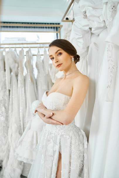 A young brunette bride stands among a rack of dresses in a wedding salon, choosing her perfect gown. - Фото, зображення