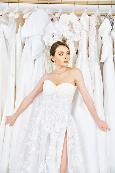A young brunette bride stands in a wedding salon, surrounded by a rack of beautiful dresses, contemplating her options. - Фото, изображение