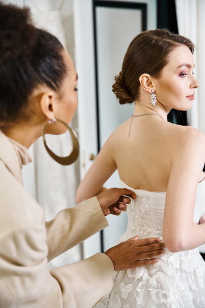 Een jonge brunette bruid in een witte jurk helpen van een vrouw zet oorbellen in een bruiloft salon. - Foto, afbeelding